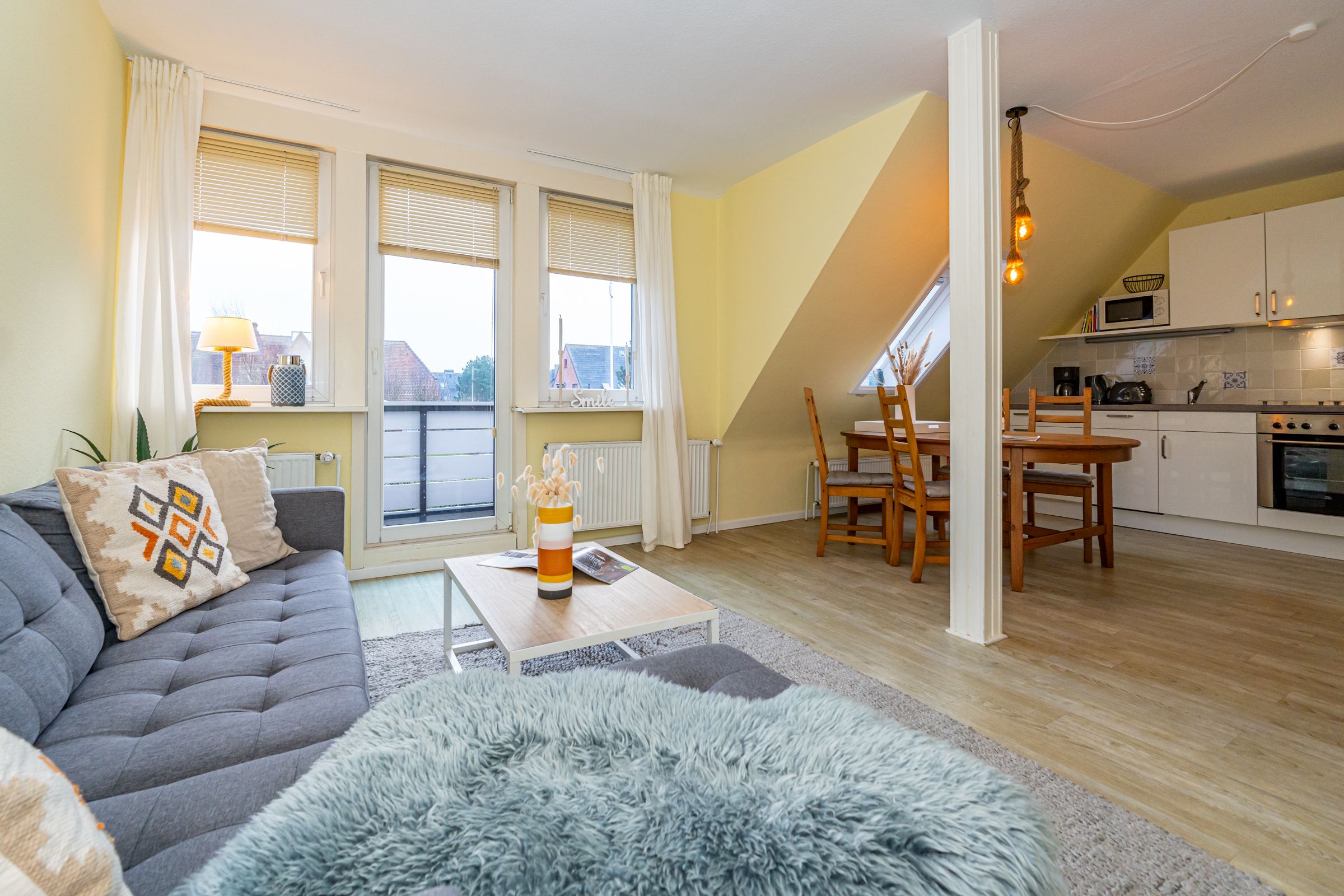 Wohnzimmer Sofa und Wohnzimmertisch mit Dekorationsartikeln und Blick auf die Balkontür und Fenster. Im Hintergrund: Essbereich und Küche