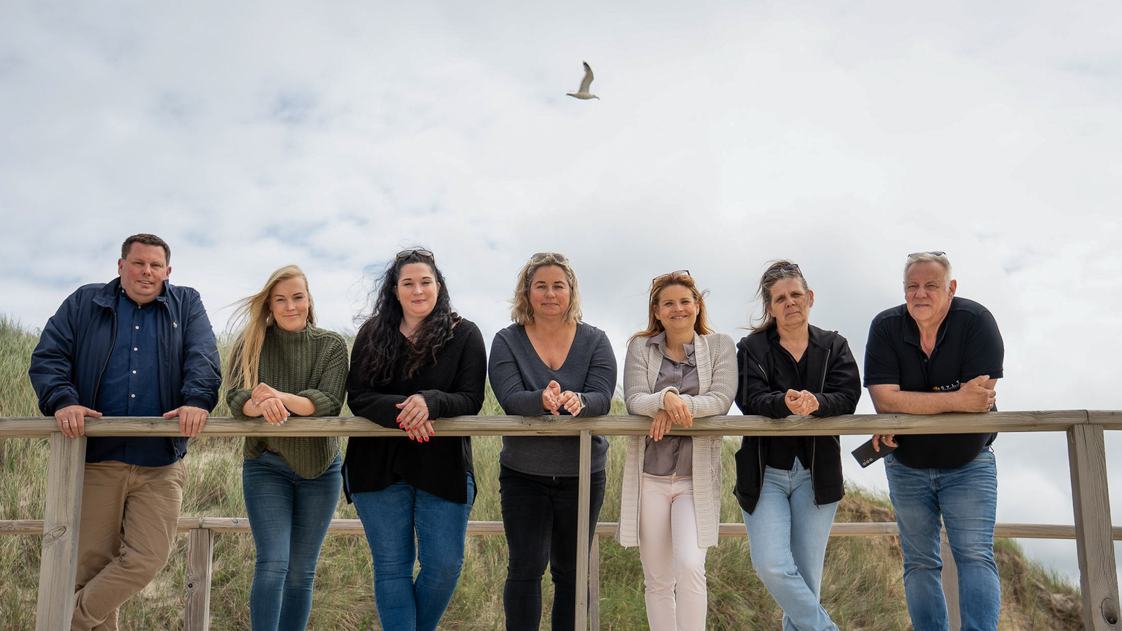 7 Menschen, zwei Männer, fünf Frauen auf einem Holzsteg. Im Hintergrund eine Düne